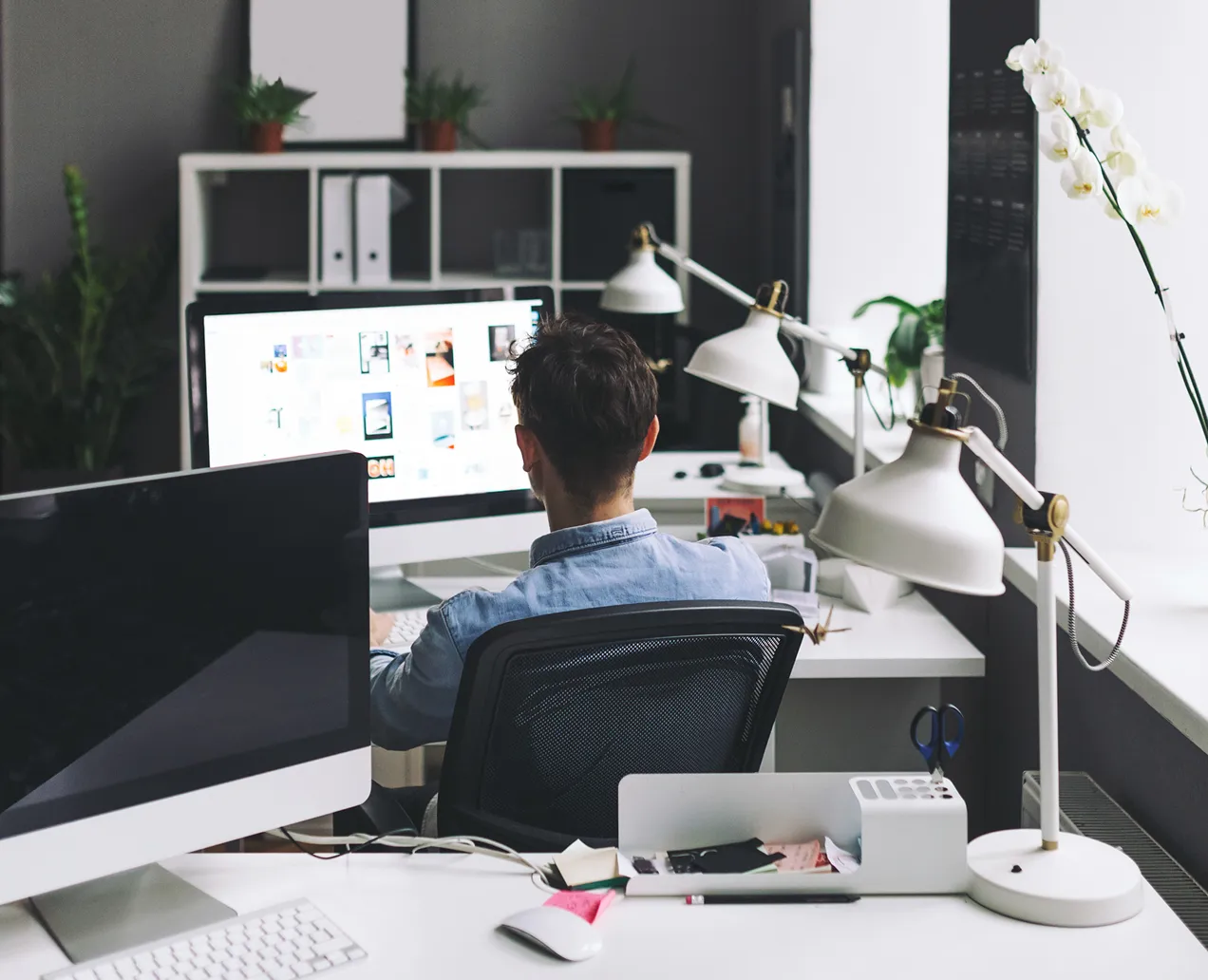 Person seated at a desk working on a computer with a bright screen in a modern office space featuring desk lamps, plants, and office supplies.